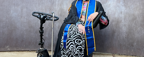 Natalee is a white person with straight dark brown hair wearing a black cap and gown and blue and gold UCLA sash, a colorful patterned long sleeve mesh top and a black and white swirl patterned skirt. They are seated on a black mobility scooter next to a silver quad cane outside in front of a rusted metal Richard Serra sculpture. They have their legs crossed and are smiling.