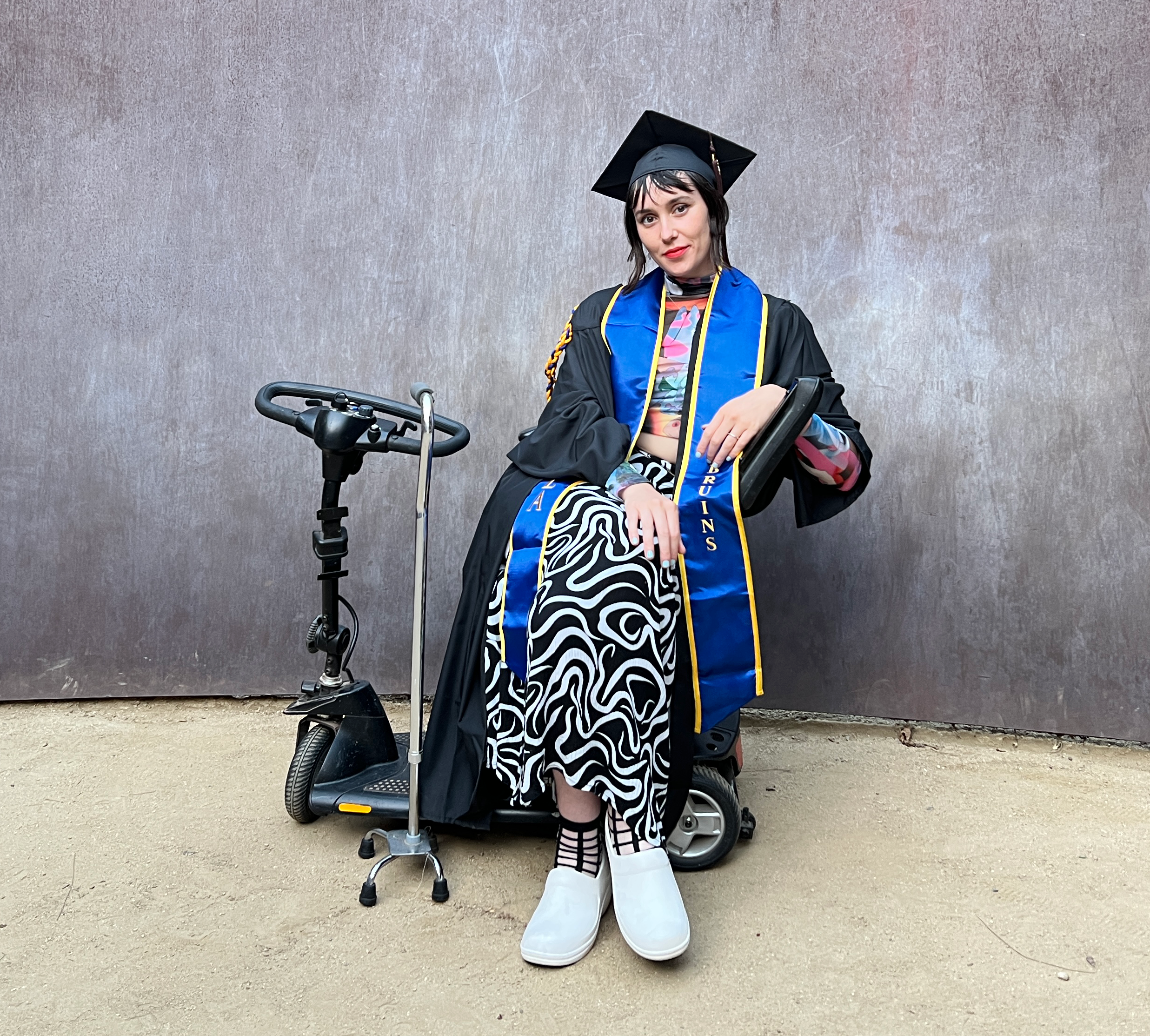 Natalee is a white person with straight dark brown hair wearing a black cap and gown and blue and gold UCLA sash, a colorful patterned long sleeve mesh top and a black and white swirl patterned skirt. They are seated on a black mobility scooter next to a silver quad cane outside in front of a rusted metal Richard Serra sculpture. They have their legs crossed and are smiling.