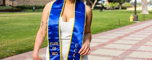 Quinn, a mixed race person with short black hair and some tattoos visible on her arms smiling at the camera. She is wearing silver hoop earrings and a white dress with a "UCLA Class of 2022" graduation sash. They are standing in front of Powell Library on campus in black ankle boots.