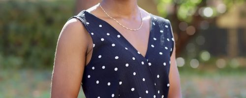 Photo of Cheri Sledge. Cheri wearing a Black and white polka dot dress. She is standing in front of green bushes and her arms are crossed over her stomach. Cheri's hair is tucked back into a low bun and she is smiling.She has a gold necklace around her neck and a purple watch on her right wrist.