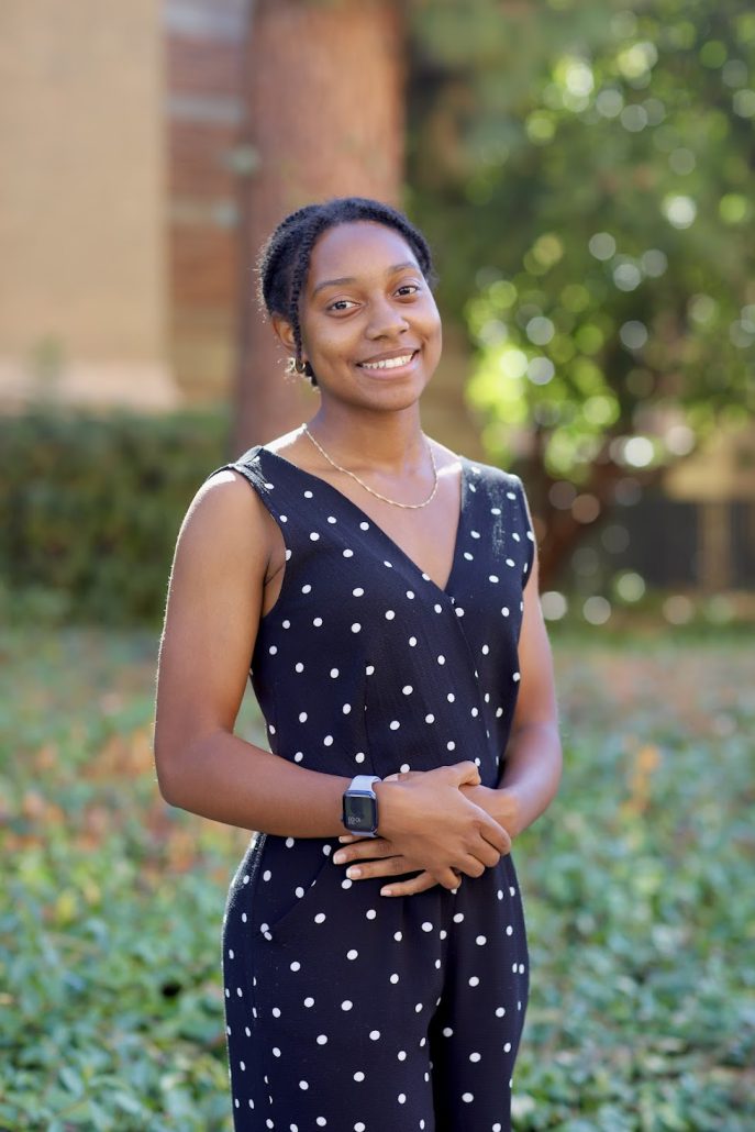Photo of Cheri Sledge. Cheri wearing a Black and white polka dot dress. She is standing in front of green bushes and her arms are crossed over her stomach. Cheri's hair is tucked back into a low bun and she is smiling. She has a gold necklace around her neck and a purple watch on her right wrist.