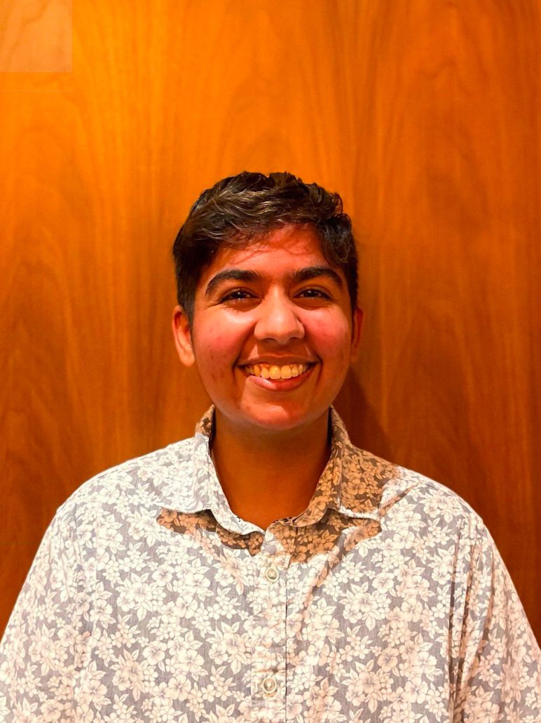 An image of Vinita from the shoulders up in front of a brown wooden wall. They are an Indian person with short black hair. They are smiling at the camera while wearing a gray and white floral patterned button-up shirt.