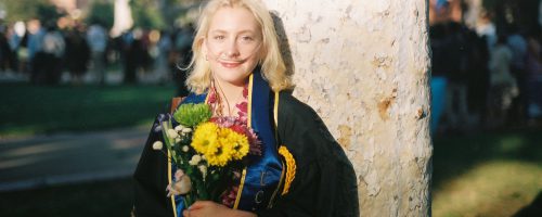 Rowan, a white woman with shoulder length blonde hair, wearing UCLA graduation gown and sash, and using her supplemental oxygen. They are holding a bouquet of flowers and standing in front of a tree in Dickson court at UCLA. She is smiling at the camera.