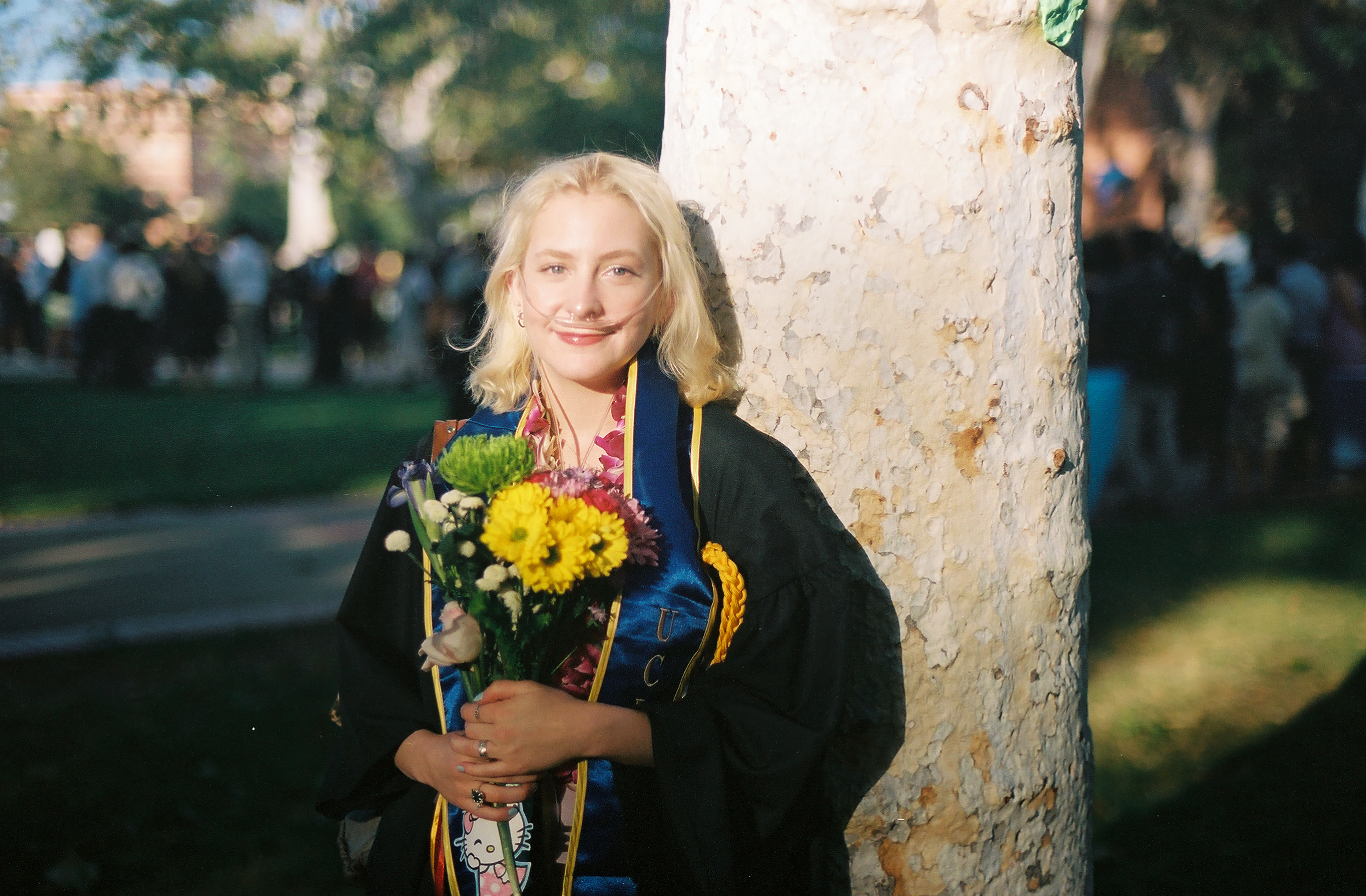 Rowan, a white woman with shoulder length blonde hair, wearing UCLA graduation gown and sash, and using her supplemental oxygen. They are holding a bouquet of flowers and standing in front of a tree in Dickson court at UCLA. She is smiling at the camera.