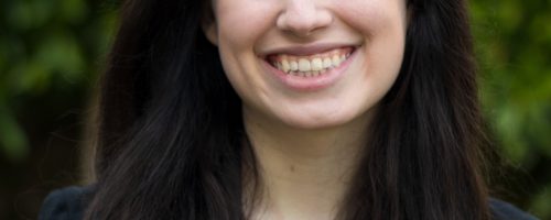 A photo of Rachel Rothschild smiling from the chest up standing in front of a mossy wall and trees. She is wearing a tan shirt with a blazer and has light skin and long brown hair.