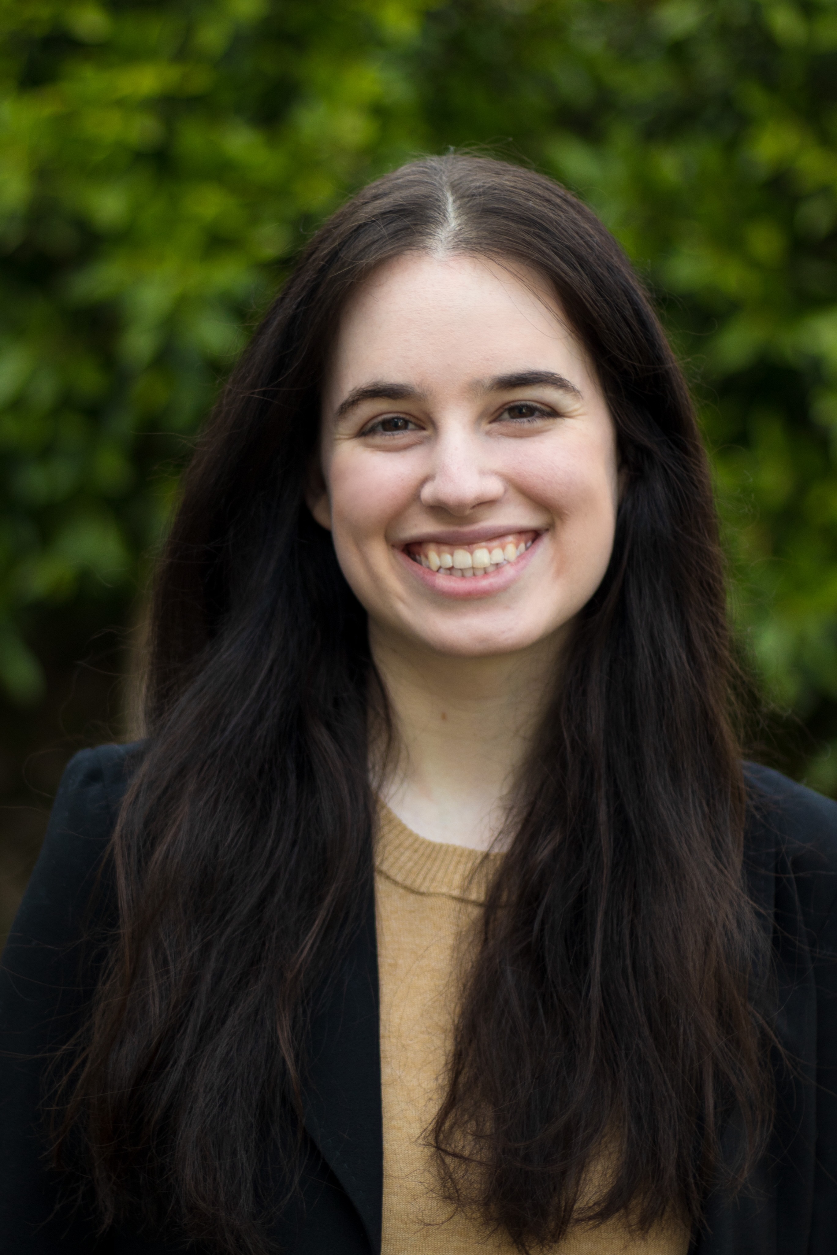 A photo of Rachel Rothschild smiling from the chest up standing in front of a mossy wall and trees. She is wearing a tan shirt with a blazer and has light skin and long brown hair.