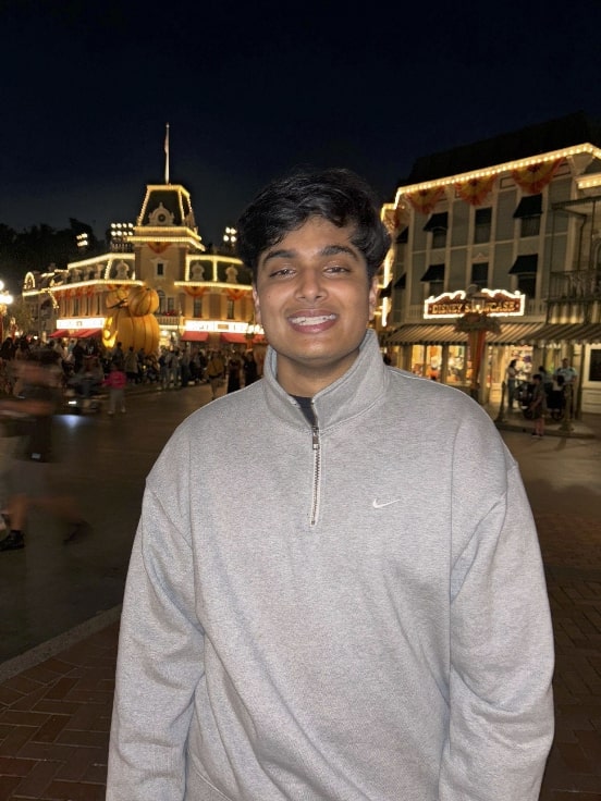 Aryan, a young man with short black hair, smiles in front of an amusement park. He is wearing a gray sweatshirt.