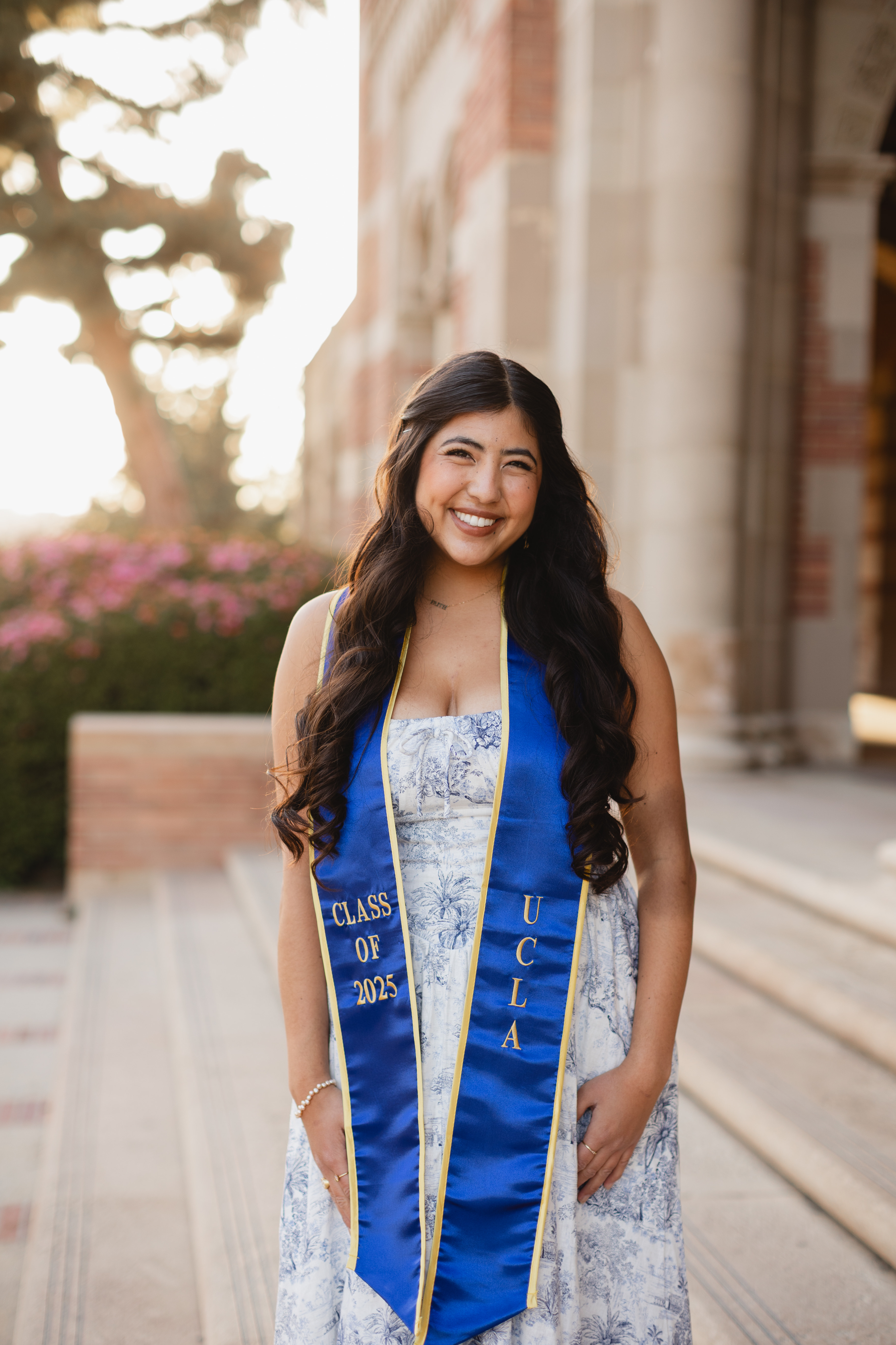 Lani, a young woman with long, dark brown hair poses in front Royce Hall. She is wearing a white and blue dress with a blue and gold 