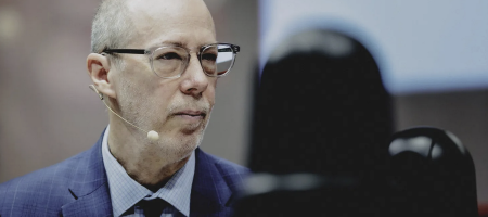 Jack Bobo wears a blue suit and glasses. He looks to the side as he listens to the speaker.