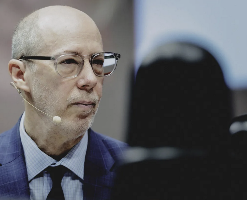 Jack Bobo wears a blue suit and glasses. He looks to the side as he listens to the speaker.