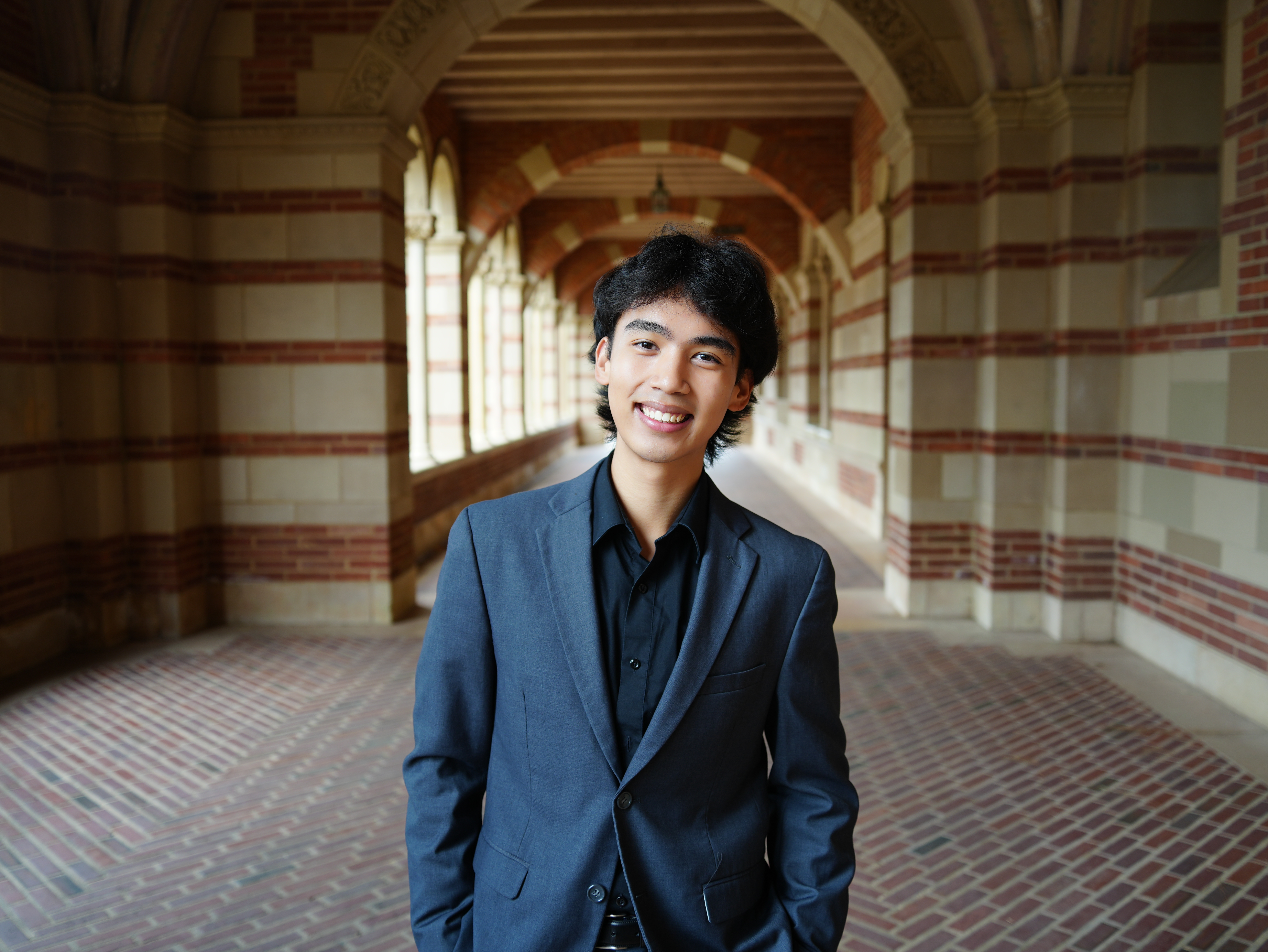 Elijah Bautista, a Filipino-American man with black hair, is pictured wearing a dark blue blazer over a black button-up shirt. He stands smiling at the camera with his hands at his sides. The photo was taken at UCLA’s Royce Hall, where he is positioned beneath a walkway framed by brick and stone arches with pillars along the sides.