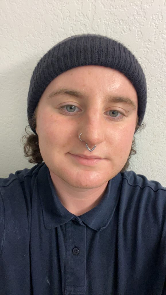 Close-up photo of Roxas, a white-skinned nonbinary person with freckles on their face, a triangle purple-jeweled septum ring, and a silver nostril ring. They have blue eyes and a black beanie with medium-length brown hair peering through. They are also wearing a dark blue collared shirt, and are positioned against a white textured wall.