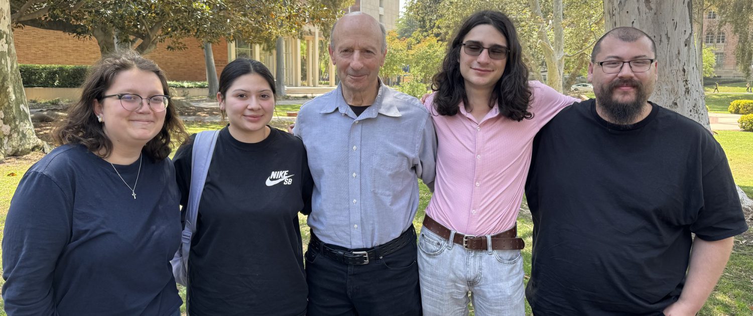 Pictured from left to right is Isabella Latcham-Radusky, Natalya Venegas, Professor William Gelbart, Marcy Mazin and Emil Dominguez. They are standing with their arms on each other's shoulders in front of trees and greenery.