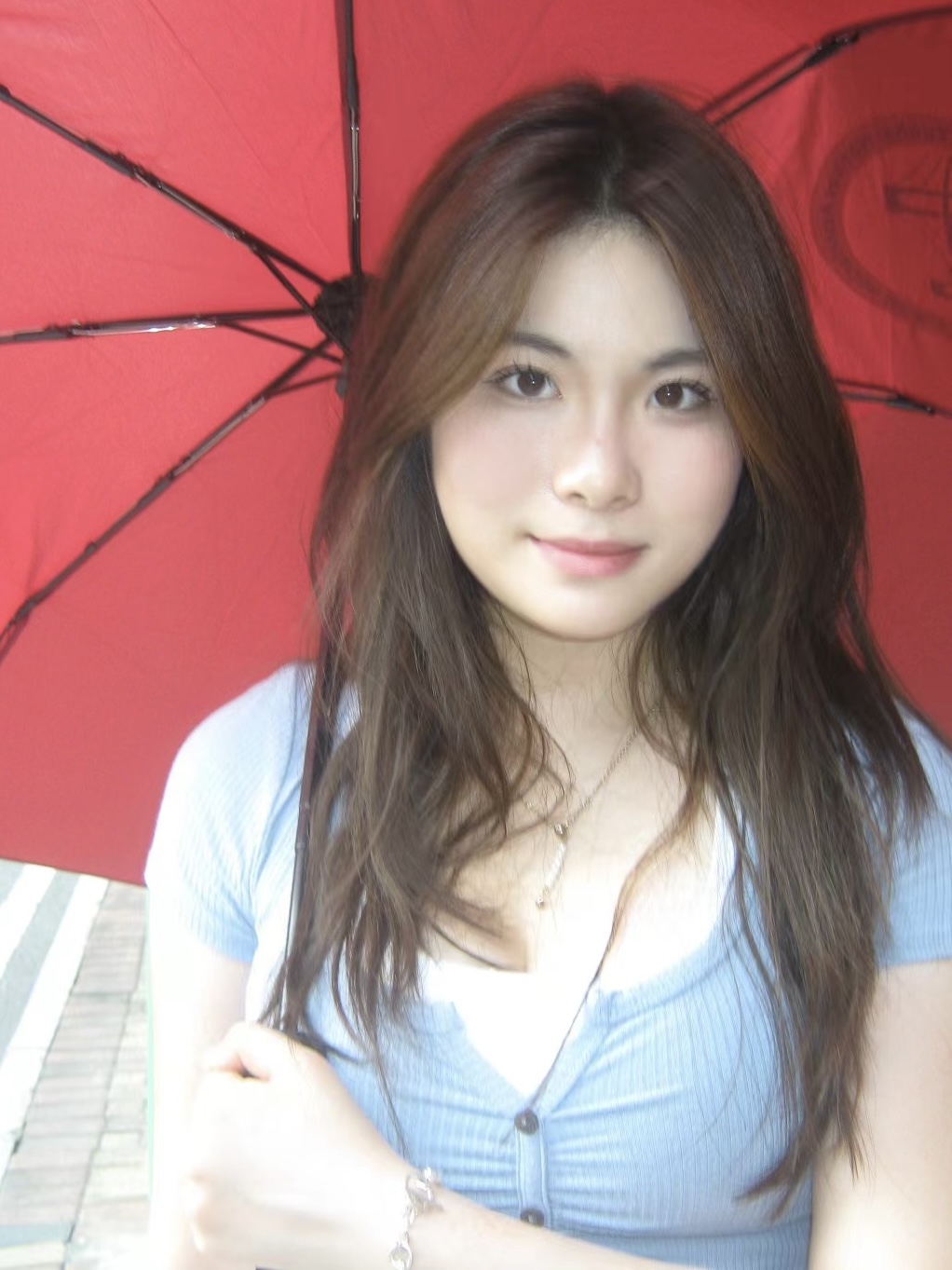 Photo of Chiu Yu Lumina Chan. She has long brown hair and brown eyes and is wearing a light blue short-sleeved top over a white camisole. Lumina is holding a red umbrella while standing on a city street.