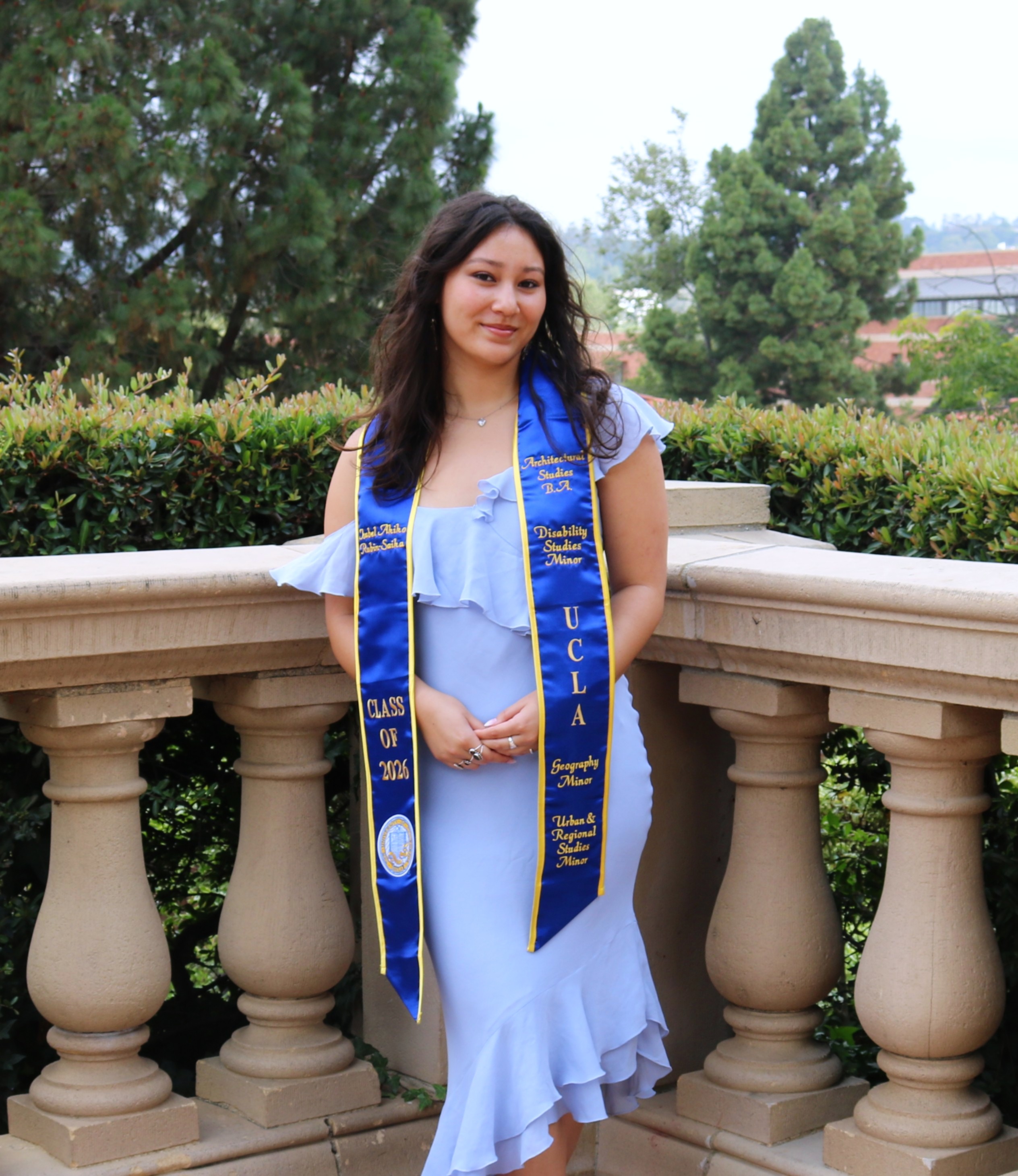 Isabel, a young woman with medium-length brown hair, stands in front of a decorative railing with foliage behind. She is wearing a blue dress with ruffles and a blue and gold 
