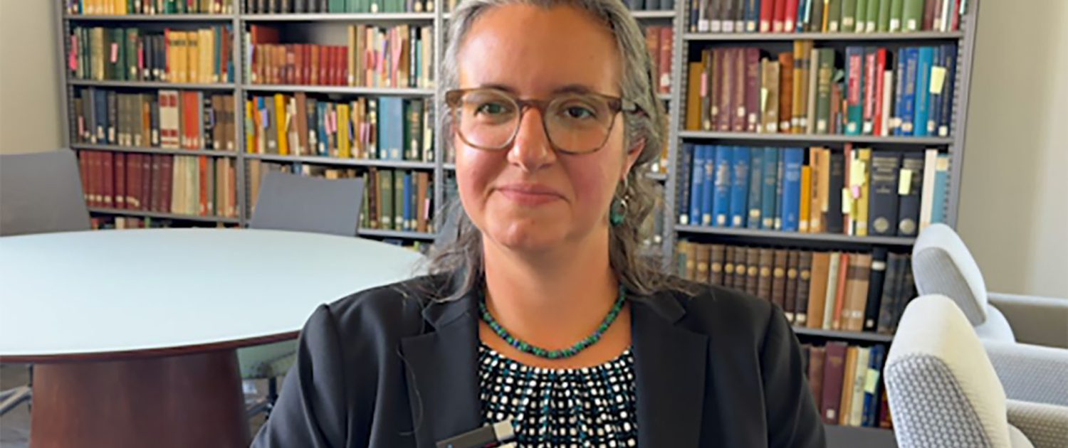 Midrange shot of Lara Fabian in dark blue jacket and patterned top, seated in a room with a bookshelf in the background.