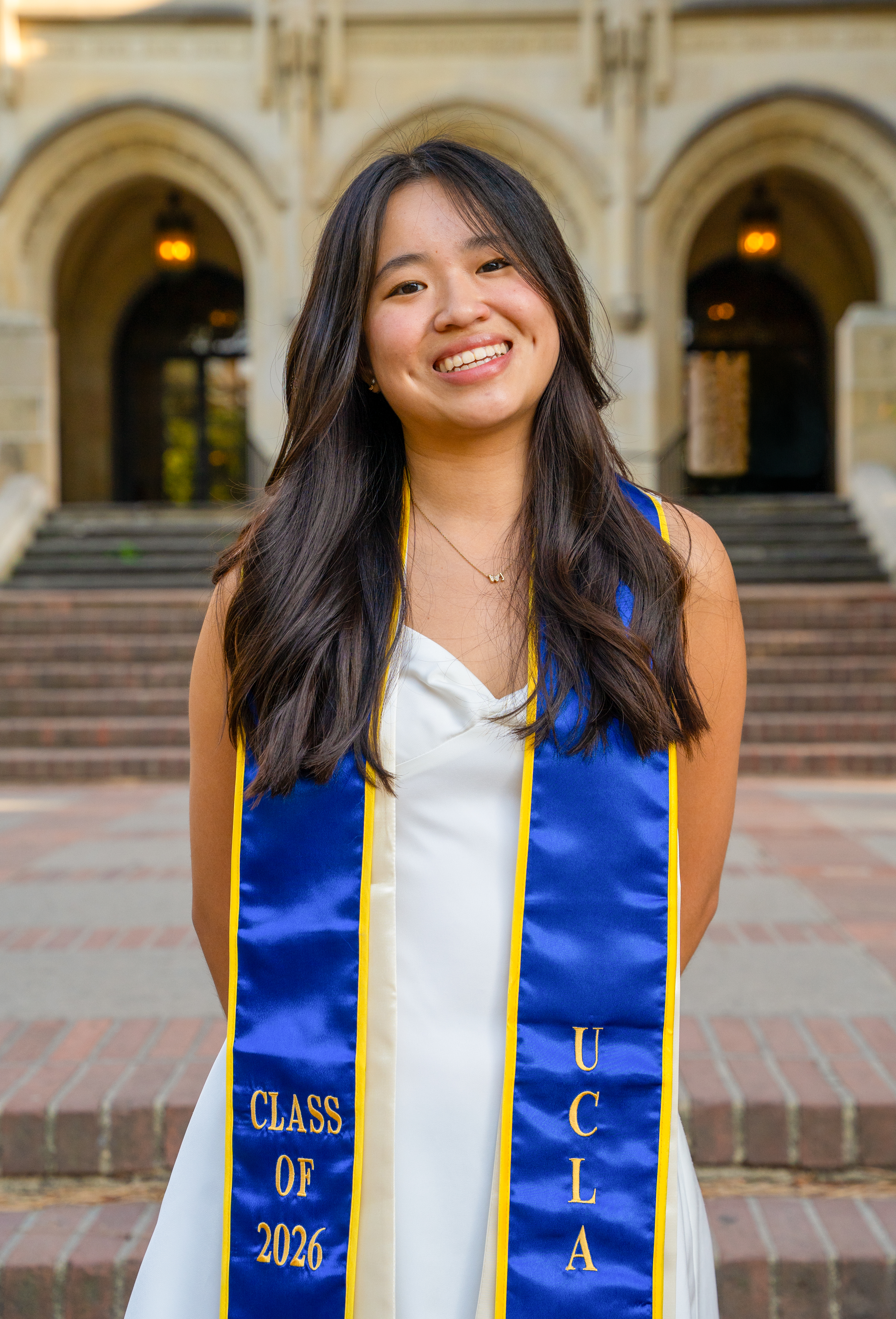 Jessica, a young woman with long, curled brown hair stands in front of Kerckhoff Hall. She is wearing a satin white dress with a blue and gold “Class of 2026” graduation stole.